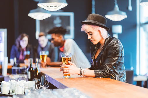 Cool blond young woman wearing leather jacet and hat drinking beer in the pub. Group of friends in the background.