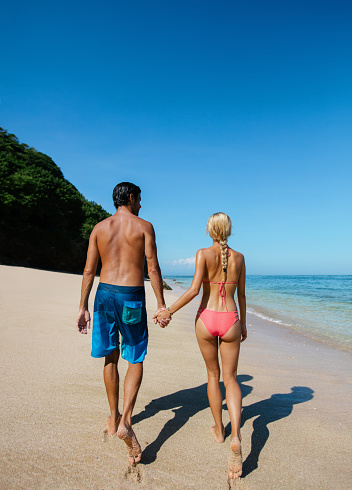 Rear view shot of young couple holding hands walking along the beach. Couple in love on summer holidays on tropical beach.