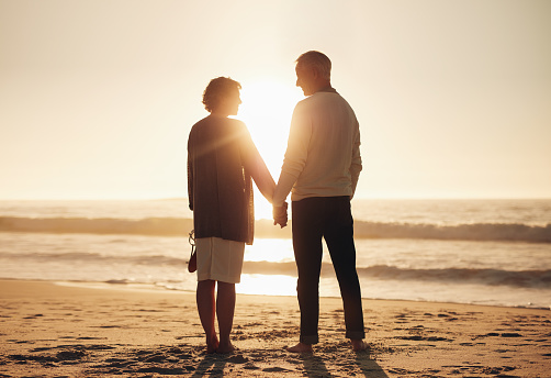 Rear view of a senior couple holding hands on the beach. Mature couple standing together on a seashore at sunset.