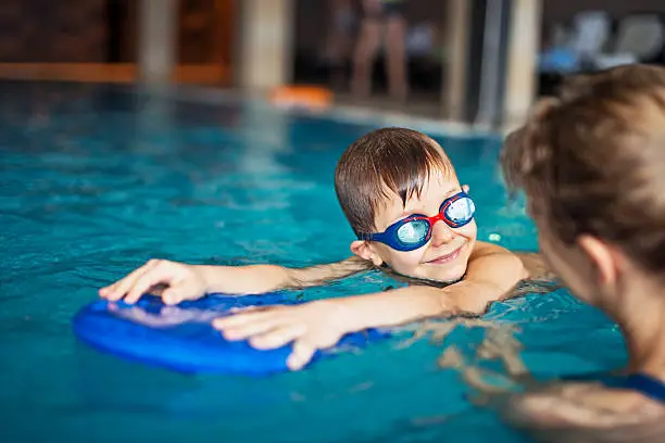 Happy little boy during swimming lesson smiles at his mother. The boy is aged 6 and is wearing swimming goggles. The boy is using a kickboard.