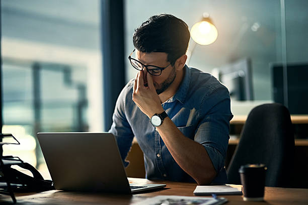 Burnout is killing his career Shot of a young businessman experiencing stress during a late night at work frustration stock pictures, royalty-free photos & images