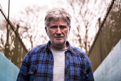 Horizontal close up image of a homeless senior man of caucasian ethnicity looking at the camera. He is outdoors in winter, and wears an old batter blue check lumberjack jacket. His grey hair is unruly and unwashed. The man's facial expression is one of sadness and despair. Horizontal colour image with copy space.