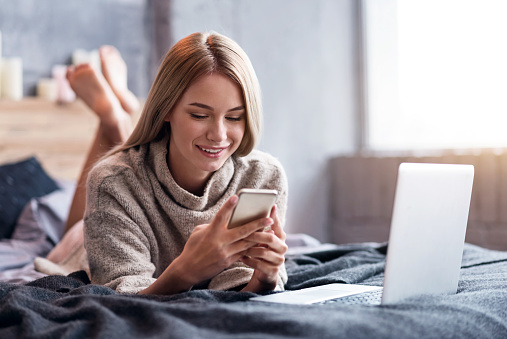 Technologies and rest. Delighted pretty young woman using smartphone and laptop while lying in bed and relaxing.