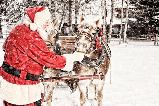 Outdoor Snowing Scene.  Santa in his red suit feeding a team of horses a green apple.  There is a cowboy in the background.  He appears to be waiting for Santa so they can start moving the team out on Christmas Eve.