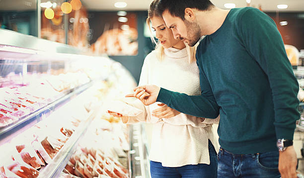 Couple buying fresh meat in supermarket. Closeup side view of a late 20's couple choosing some fresh meat for tonight's dinner. They are reading label on one of the packages and seem a bit uncertain about it. meat locker photos stock pictures, royalty-free photos & images