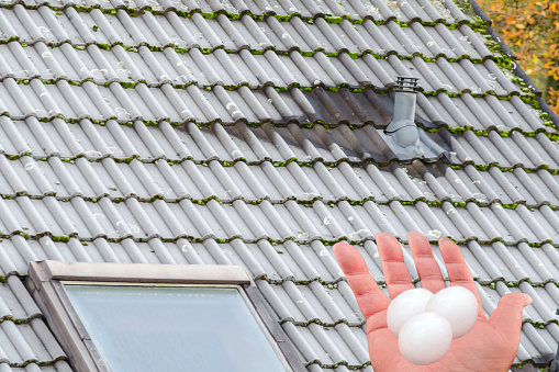Severe damage to the roof by hail. Human hand with hail grains in front of a building roof.