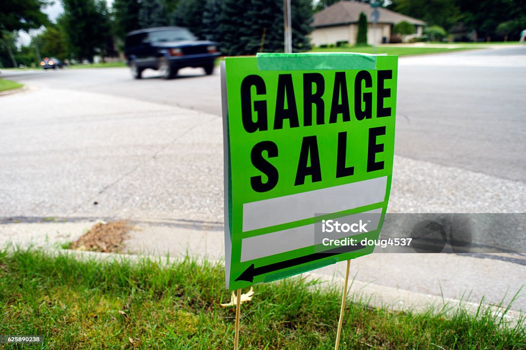 Garage Sale Sign Next to Neighborhood Street A green Garage Sale sign placed in the grass next to the curb, pointing to where the sale is held.  The image was made with a wide angle lens giving context as to where the sign is placed and a blurred car is on the street in the background. Advertisement Stock Photo