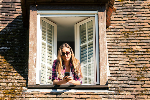 Young woman showing throuth the window and using smart phone. Wears checkered shirt and white top, sunglasses. Old architecture house-top with opened vintage window.
