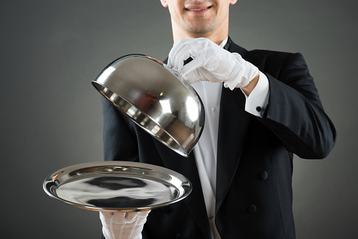 Midsection of waiter holding cloche over empty tray while standing against gray background
