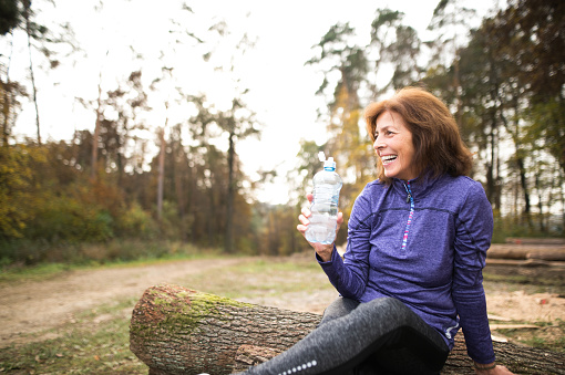 Senior runner in nature. Woman sitting on wooden logs, resting, holding water bottle.