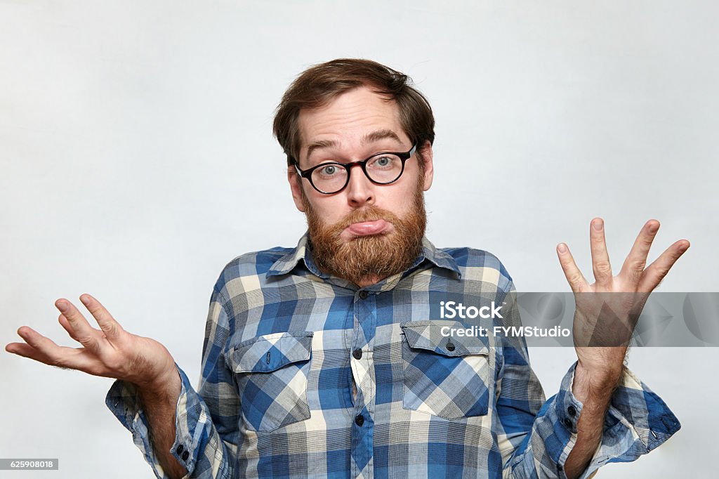 Confused bearded man in eyeglasses shrugging his shoulders Man with beard and eyeglasses shrugging his shoulders in confusion, arms raised, isolated on gray Confusion Stock Photo