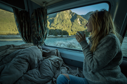 Young woman in a camper van drinks a tea and looks through window. Beautiful mountain landscape in early morning, Autumn/Winter season.