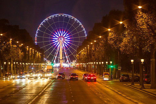 A night shot of Champs-Élysées Avenue. A short row of cars is visible in foreground. La Grande Roue de Paris (Paris Big Wheel), at Place de la Concorde, can also be seen In the background.