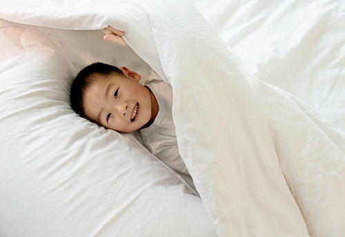 Smiling little boy hiding under blanket in bed.