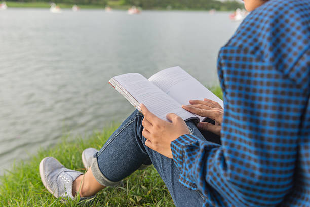 istock Young girl sitting on grass reading book beside lake 625940632