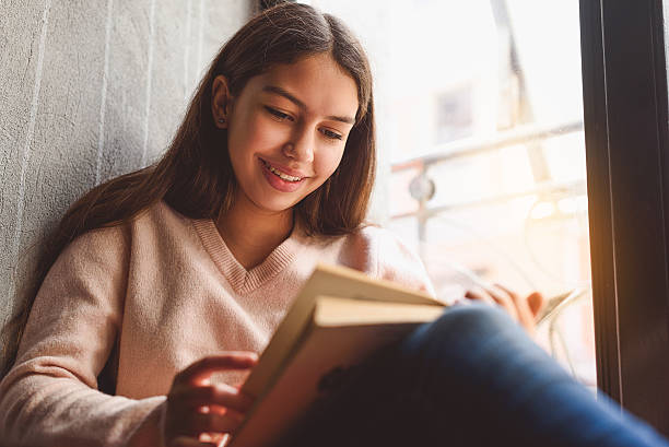 Girl absorbedly reading open book Smiling female teenager is on sill, holding disclosed volume poetry literature photos stock pictures, royalty-free photos & images