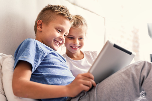 Growing up with technology. Two little smiling kids sitting on bed at home and playing games on their technology tablet