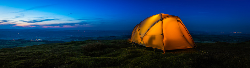 Warmly illuminated dome tent pitched on a picturesque mountain top wild camp site overlooking lights in the valley far below at sunset.