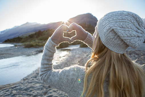 Young woman by the lake makes a heart shape finger frame. Lake and mountain landscape on the background. 