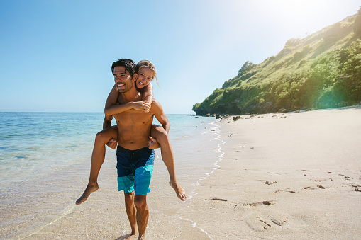 Happy couple in love on beach summer vacations. Joyful woman piggybacking on boyfriend, playing and having fun on travel vacation.
