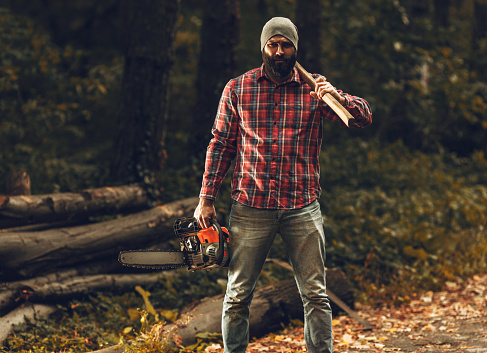 Lumberjack worker standing in the forest with axe and chainsaw