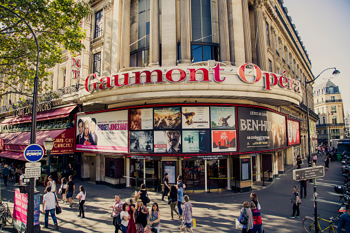 Paris, France - September 9, 2016: The Cinema Complex Gaumont Opera, on rue des Capucines in the 9th Arrondissement. Its construction took 2 years, from 1866 to 1868. Originally used for Vaudeville theatre, its vocation changed to cinema in 1927 and was then called Paramount Opera. Europalaces bought it in 2007 and since then, this Cinema Complex bears the name Gaumont Opéra. It's neighbour, Le Grand Café des Capucines, and some people are also visible in the image.
