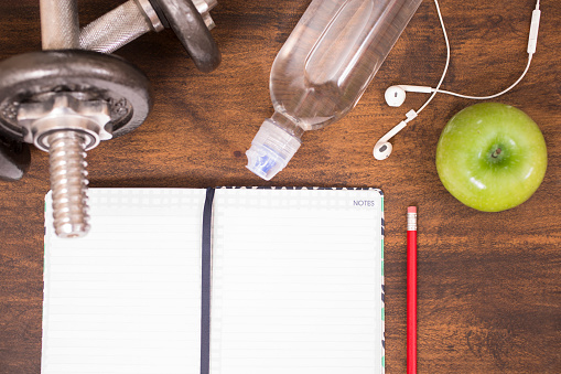 Fitness, exercise themed scene with barbells, notebook, earbuds, water bottle, pencil, and green apple.  Group of objects lie on wooden surface in gym or health club setting.  High angle view. Copyspace on blank journal.