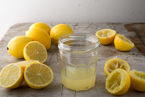 A jar of lemon juice surrounded by whole, sliced and squeezed lemons on a wooden counter.