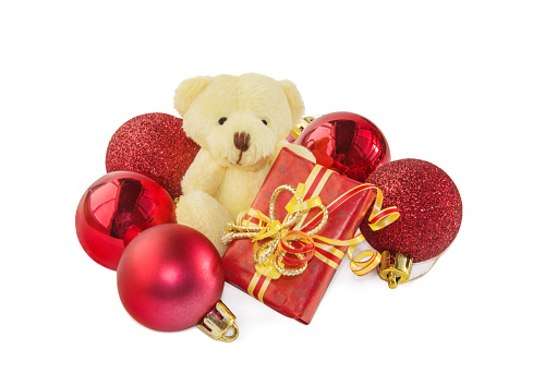 Teddy bear, classic soft toy sitting with gift box and surrounded by red Christmas balls isolated over white. Front view. Christmas and New Year theme.