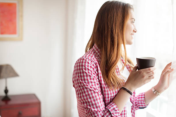 istock Smiling young woman drinking coffee and looking through window 626090518