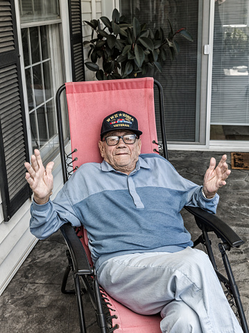 A proud, real person, 93 year old senior adult man United States Army World War II and Korean Conflict US Air Force military veteran at home relaxing on his front porch. He is shrugging his shoulders with his hands outstretched in exaggerated innocence as he listens during a conversation with a visitor. He's wearing a common, unbranded, generic souvenir shop military veteran commemorative baseball style cap with generic wording, replica plastic insignia pins and replica campaign ribbon iron-on patches.