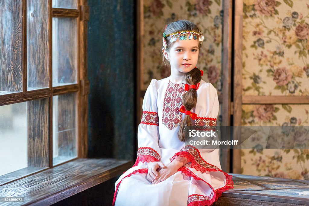 Nice Girl In Folk Costume Beautiful elementary age girl with a long braid. The girl is dressed in Mari native dress and headdress made of beads and coins on her head. She is sitting on the old trunk near a big window. The little girl is smiling looking at the camera. Studio shooting Arts Culture and Entertainment Stock Photo