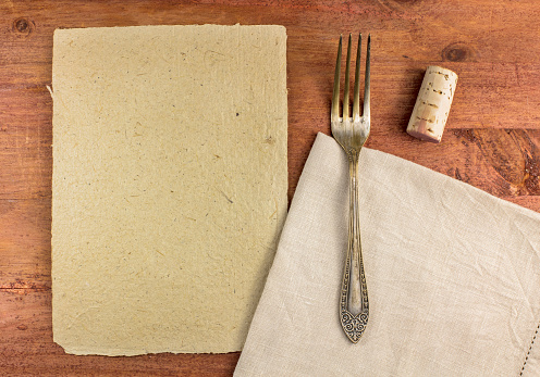 An overhead photo of a piece of parchment for copyspace, with a vintage fork and a cork, on a textile napkin. A restaurant menu or special offer banner design template