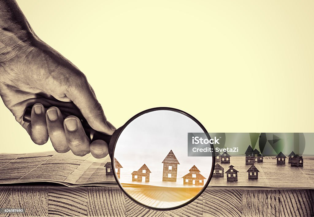 Paper house under a magnifying lens Hand holds the magnifying glass in front of an open newspaper with paper houses. That could mean rent, search, purchase real estate. Real Estate Stock Photo