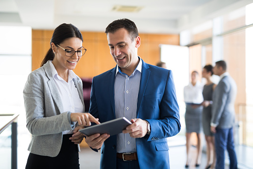 Two mixed-age business people are discussing business strategy in the office building hallway using digital tablet. There are people in the background communicating. Copy space.