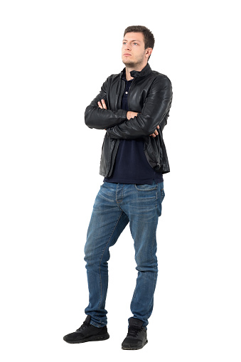 Serious young casual man in leather jacket and jeans with crossed arms looking away. Full body length portrait isolated over white studio background.