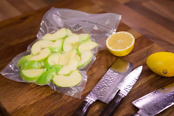 Apple in vacuumed package with lemon and knives on cutting board.