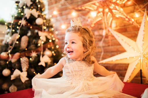 Little girl in red dress smiling by Christmas tree. Little beautiful girl in a red evening dress the Christmas tree. Girl in red in Christmas decorations. Christmas concept. New Year. Baby near a Christmas tree. High quality photo.