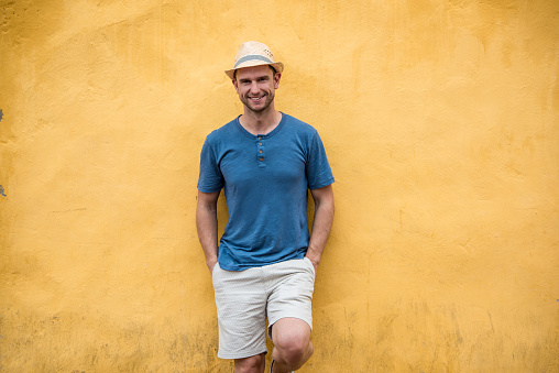 Casual male tourist looking very happy leaning against a yellow wall and smiling - people traveling concepts