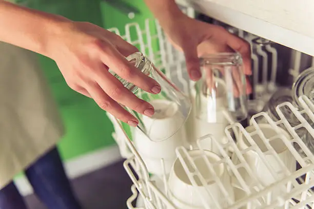 Cropped image of beautiful young woman putting dishes into dishwasher in kitchen