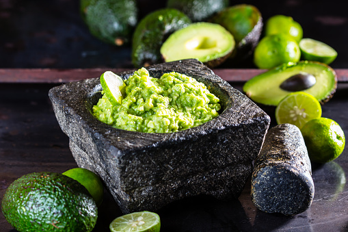 Latinamerican mexican sauce guacamole with avocado and lime in stone mortar and ingredients - fresh avocado palta, lime on black background.