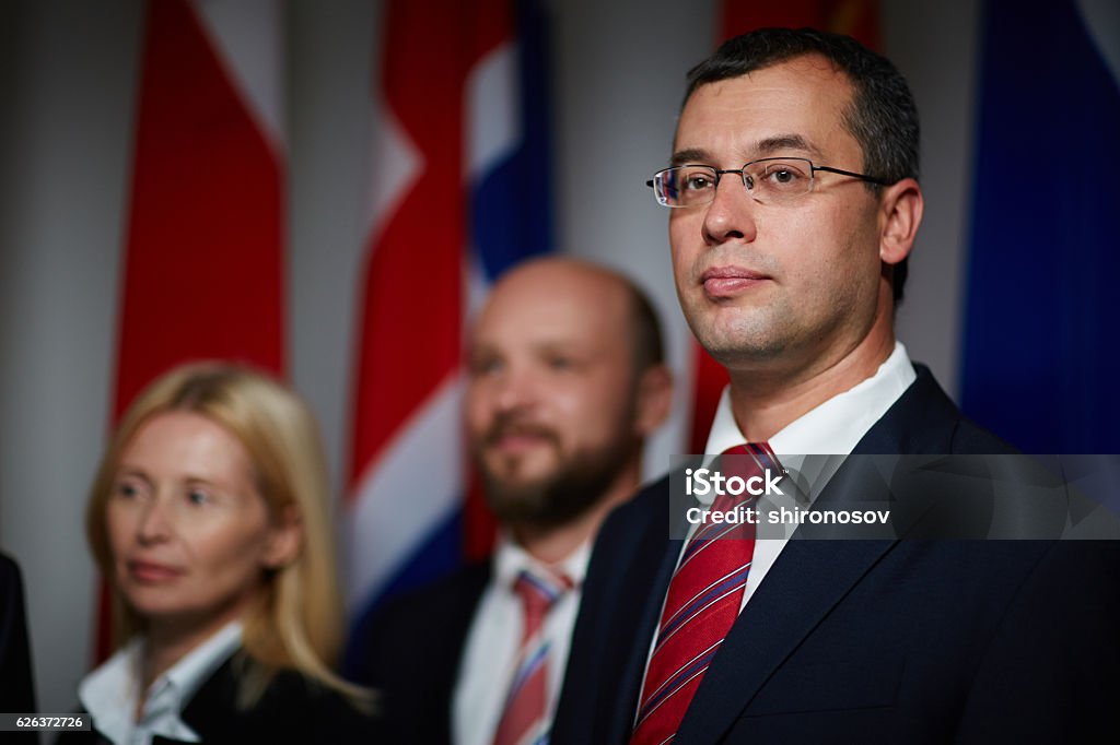 Confident good-looking politician Close-up portrait of confident, attractive, middle-aged politician wearing glasses with his colleagues and national flags on blurred background Adult Stock Photo