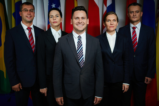 Group portrait of formal-dressed male and female delegates with confident smiling executive on foreground looking at camera on background of national flags