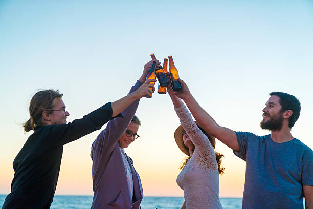 Young group of adult friends toasting using beer bottles on the beach as the sun sets. Los Angeles, America. October 2016