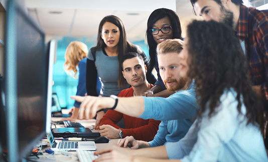 Closeup of group of application developers discussing about certain code for an application. There are three women and two men gathered around a desktop computer. They are aged from mid 20's to mid 30's, mixed race, dressed smart casually. One of the guys is pointing at the software code on the screen. Blurry people in background, also released.