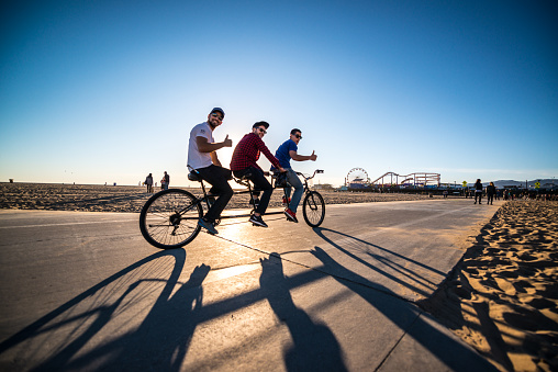 Santa Monica, USA  - January 25, 2016: People cycling  on Santa Monica beach, CA, USA. Three young men on tanned bicycle, looking at camera with thumbs up. People on background walking