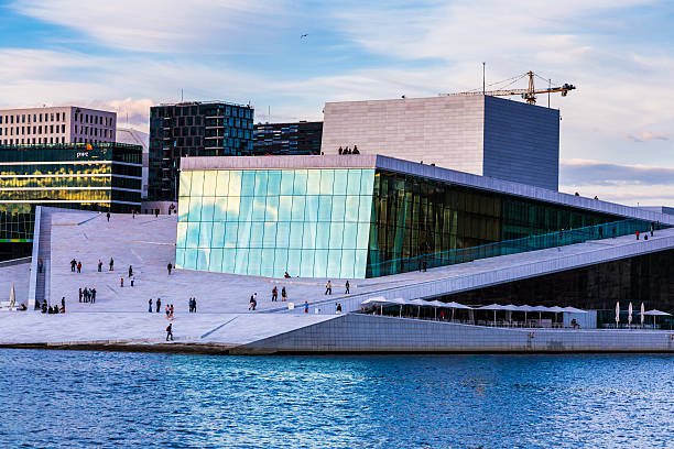 Oslo, Norway - June 12, 2015: the modern architecture of the Oslo Opera House in Oslo, Norway. Tourists walk on the marbled paving that surrounds the opera house. In the background is Oslo's modern business district, made up of multiple modern skyscrapers.