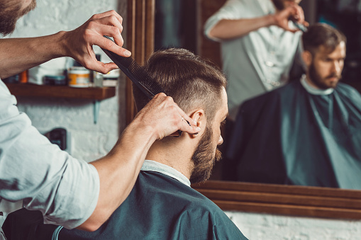 The hands of young barber making haircut of attractive bearded man in barbershop