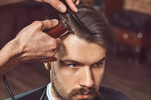 Hipster client visiting barber shop. The hands of young barber making the cut of beard