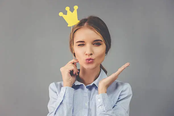 Beautiful business lady is holding party crown on stick, looking at camera and sending air kiss, on gray background